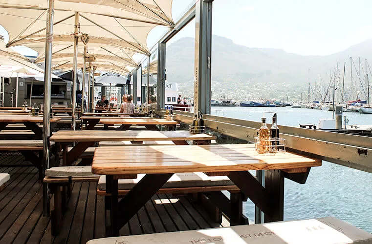 Long timber tables on the deck beside the water, mountains beyond