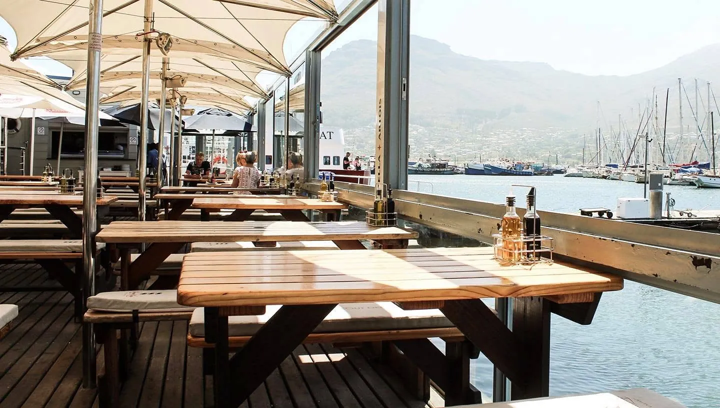 Timber tables on the deck under sail-cloth canopies, with the harbour and mountains beyond
